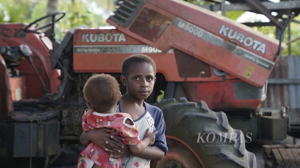 Seorang anak menggendong adiknya di depan traktor pertanian yang sudah rusak di Kampung Serapu, Distrik Semangga, Merauke, Papua, Selasa (7/11/2022).