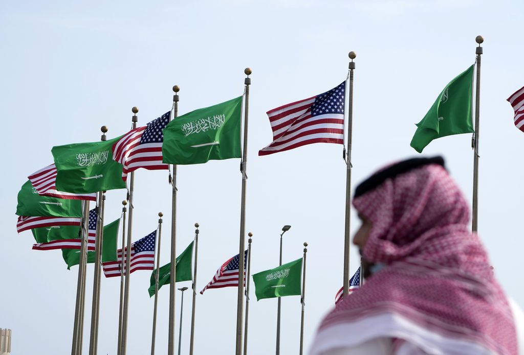 Seorang pria berdiri di dekat tiang-tiang bendera yang mengibarkan bendera Arab Saudi dan Amerika Serikat di Jeddah, Arab Saudi, Kamis (14/7/2022), menjelang kedatangan Presiden AS Joe Biden di negara itu. 