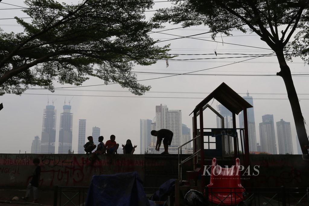 Anak-anak bermain di atas tanggul Sungai Ciliwung dengan latar belakang deretan gedung bertingkat kota Jakarta yang diselimuti kabut asap polusi, Rabu (15/6/2022). 