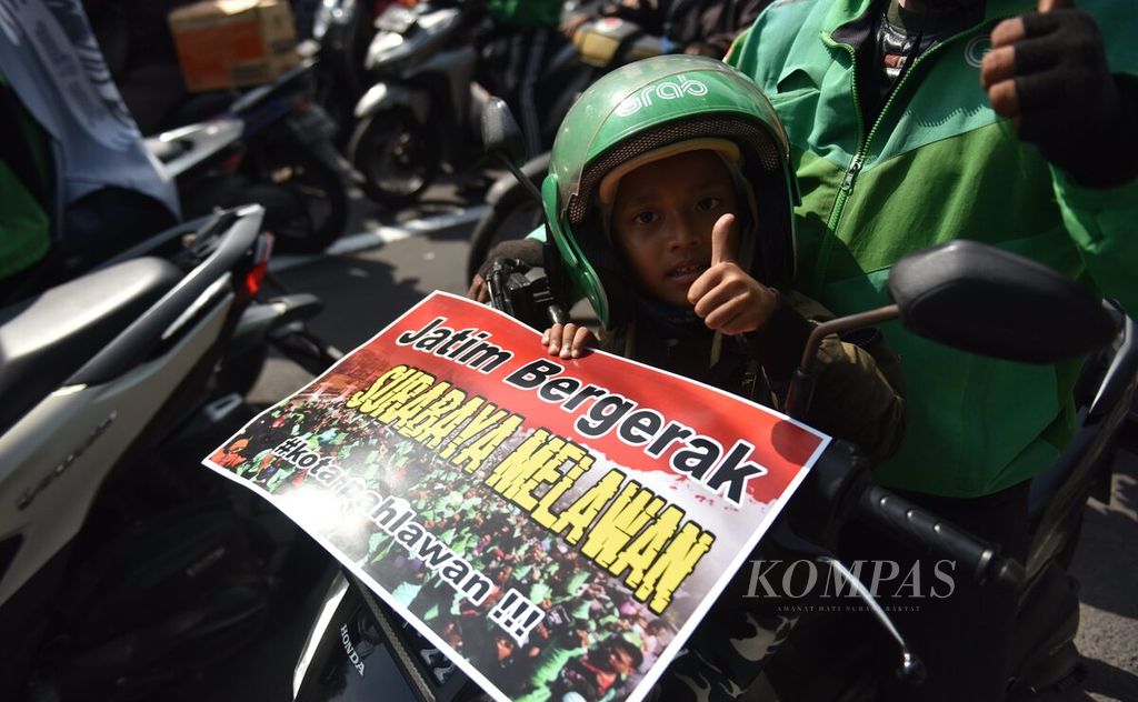 Seorang pengunjuk rasa membawa serta anaknya saat pengendara ojek daring melakukan aksi unjuk rasa di depan Kantor Dinas Perhubungan Provinsi Jatim, Surabaya, Kamis (24/3/2022). 