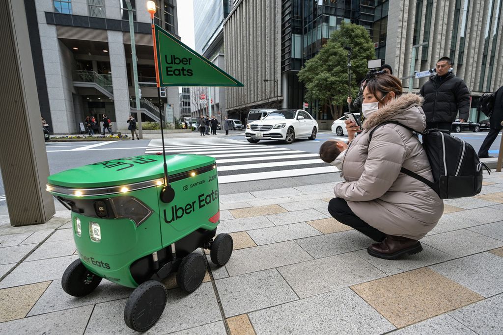 Seorang wanita menggendong bayinya mengambil foto saat robot tak berawak melewati mereka di sepanjang jalan saat ujicoba layanan pengiriman robot oleh Uber Eats Japan, Mitsubishi Electric, dan pengembang robot Cartken di pusat kota Tokyo, Jepang, 5 Maret 2024.