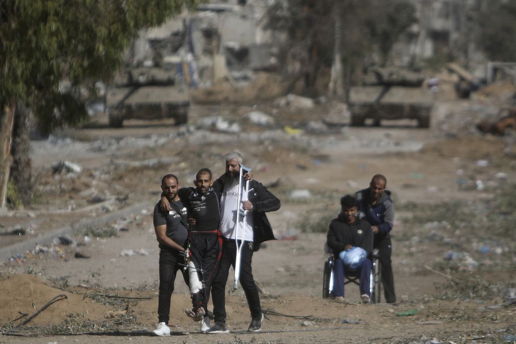 Dua orang warga Palestina, Jumat (24/11/2023) membantu warga lainnya yang kaki kanannya cedera, berjuang menuju ke Gaza selatan untuk mencari lokasi perlindungan yang lebih aman dari kemungkinan serangan Israel jika jeda pertempuran empat hari selesai. Di belakang mereka tampak dua tank milik militer Israel berjaga-jaga.. 