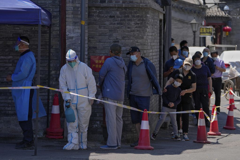 Mengenakan pakaian pelindung, seorang petugas kesehatan menyemprotkan cairan disinfektan di tengah barisan warga yang tengah mengantre untuk melakukan tes Covid-19 di Beijing, China, Senin (16/5/2022).  