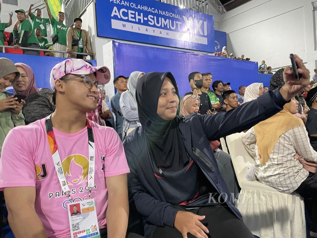 Lifter peraih medali emas Olimpiade Paris 2024, Rizki Juniansyah, melayani permintaan foto bersama dari atlet PON Aceh-Sumut 2024, Aqsha Fellisa Putri, saat berada di dalam arena angkat besi GOR Seuramoe, kompleks Stadion Harapan Bangsa, Banda Aceh, Aceh, Rabu (4/9/2024).