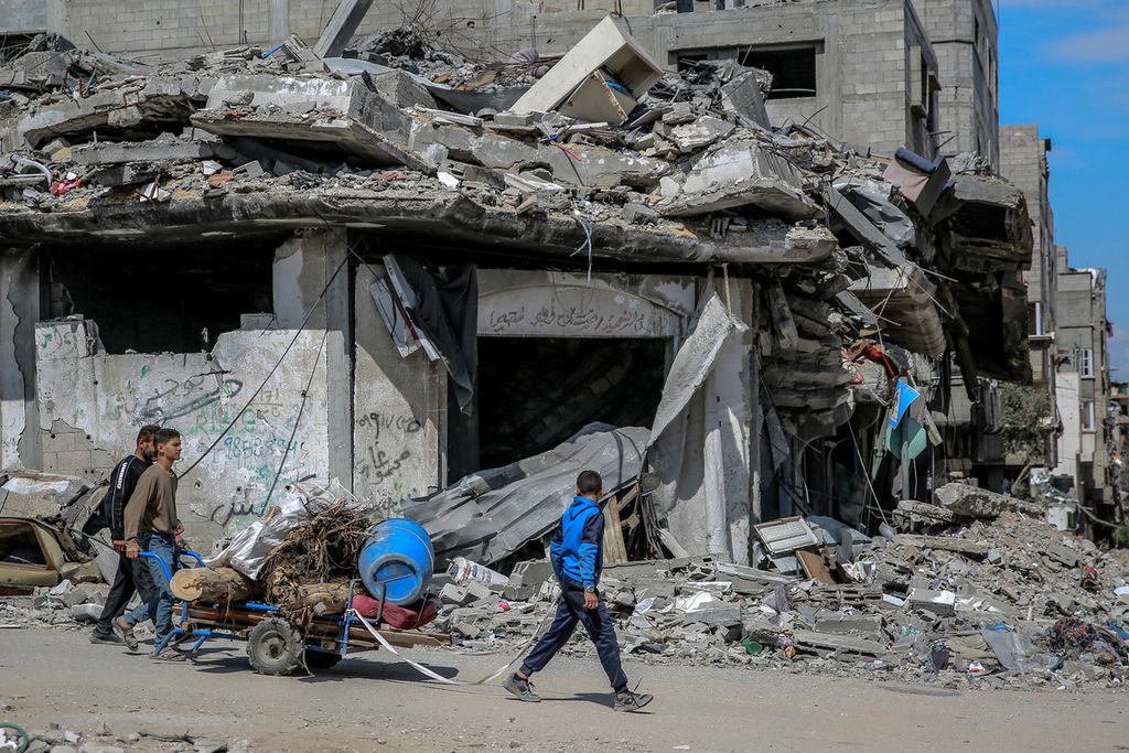 Pemuda Palestina berjalan melewati bangunan yang hancur akibat serangan Israel saat mereka mengumpulkan barang-barang yang bisa diselamatkan dari puing-puing di kota Gaza, Senin (25/3/2024).