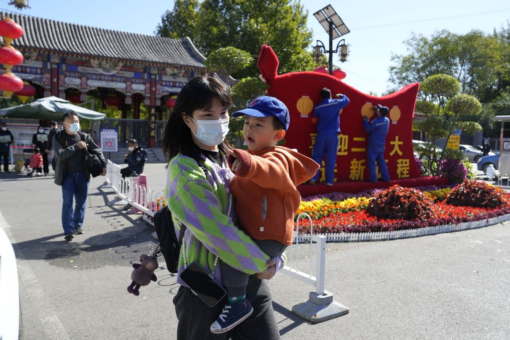 Seorang perempuan menggendong anaknya melintasi pekerja yang sedang memasang dekorasi menjelang Kongres Nasional Ke-20 Partai Komunis China di Beijing, Senin (10/10/2022). 