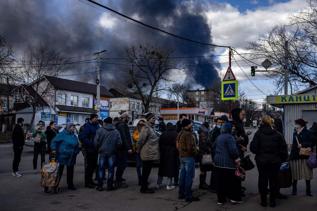 Warga mengantre di depan sebuah supermarket, sementara asap tebal membubung ke angkasa tak jauh dari mereka, di kota Vasykiv, di luar ibu kota Kiev, Ukraina, Minggu (27/2/2022).