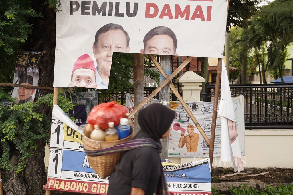 Penjual jamu keliling melintasi baliho calon legislatif dan pasangan capres-cawapres yang sobek di Jalan Pemuda, Jakarta Timur, Jumat (19/1/2024).