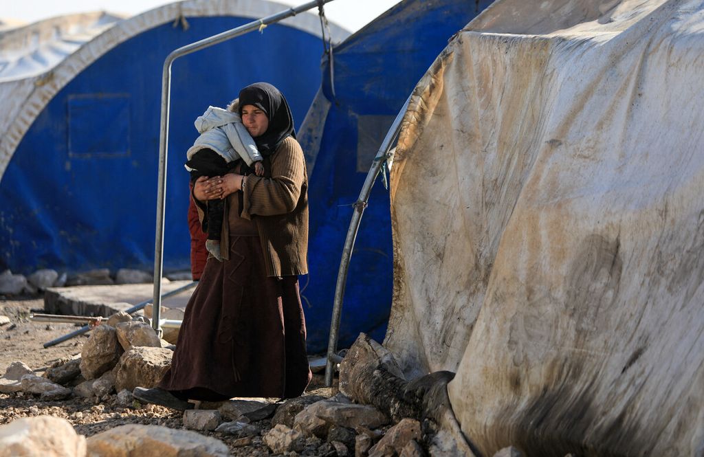 Seorang wanita pengungsi Suriah menggendong seorang anak di depan sebuah tenda di kamp sebelum direlokasi ke kompleks perumahan baru di daerah yang dikuasai oposisi Bizaah, timur kota al-Bab di provinsi Aleppo utara, dibangun dengan dukungan badan darurat Turki AFAD, Rabu (9/2/2022). 