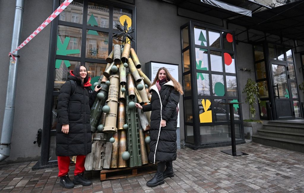 Dua perempuan berpose di samping pohon Natal simbolis yang terbuat dari cangkang kerang dan amunisi bekas lain di luar sebuah kafe di pusat kota Kyiv, Ukraina, Senin (18/12/2023).
