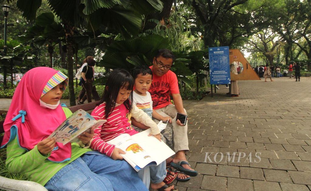 Tiga anak didampingi orangtuanya membaca buku di Taman Suropati, Menteng, DKI Jakarta, Minggu (17/7/2022). Buku itu diambil dari lemari buku Bookhive yang terletak di tengah taman tersebut.