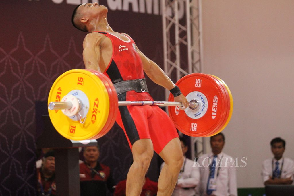 Dengan penuh kekuatan, Rizki Juniansyah, lifter muda Indonesia yang masih berusia 20 tahun, mengangkat pelat-pelat besi seusai memecahkan rekor dengan total angkatan 347 kilogram (<i>snatch </i>156 kg dan <i>clean and jerk </i>347 kg) di cabang angkat besi kelas putra 73 kilogram di National Olympic Stadium, Kamboja, Minggu (14/5/2023).