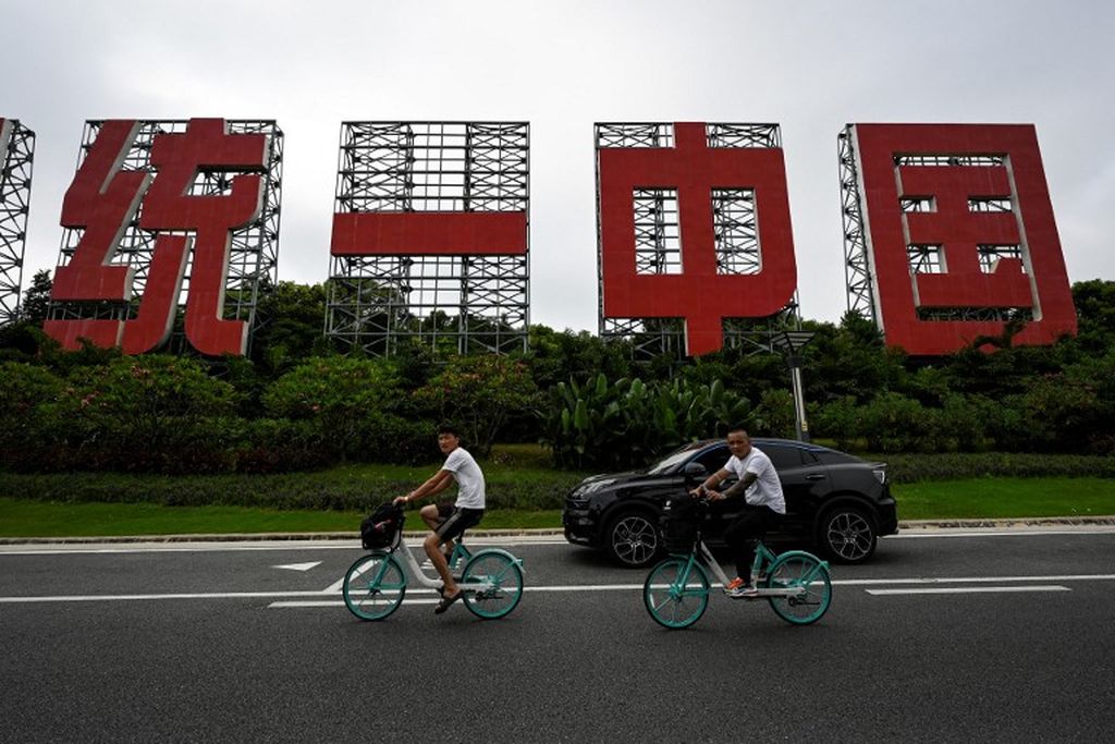 Foto yang diambil pada 25 Juli 2022 ini menunjukkan orang-orang mengendarai sepeda dan berkendara melewati sebuah papan besar bertuliskan "Satu Negara, Dua Sistem, Menyatukan China" di Xiamen, Provinsi Fujian, China.
