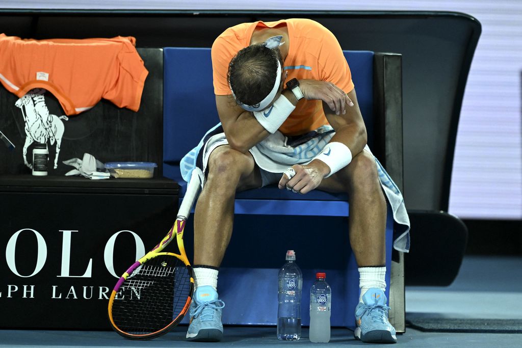 Petenis Spanyol, Rafael Nadal, tertunduk menyeka wajahnya pada jeda laga babak kedua Grand Slam Australia Terbuka melawan petenis muda Amerika Serikat, Mackenzie McDonald, di Melbourne Park, Melbourne, Rabu (18/1/2023). Nadal, yang menderita cedera pinggul pada laga itu, akhirnya kalah, 4-6, 4-6, 5-7.