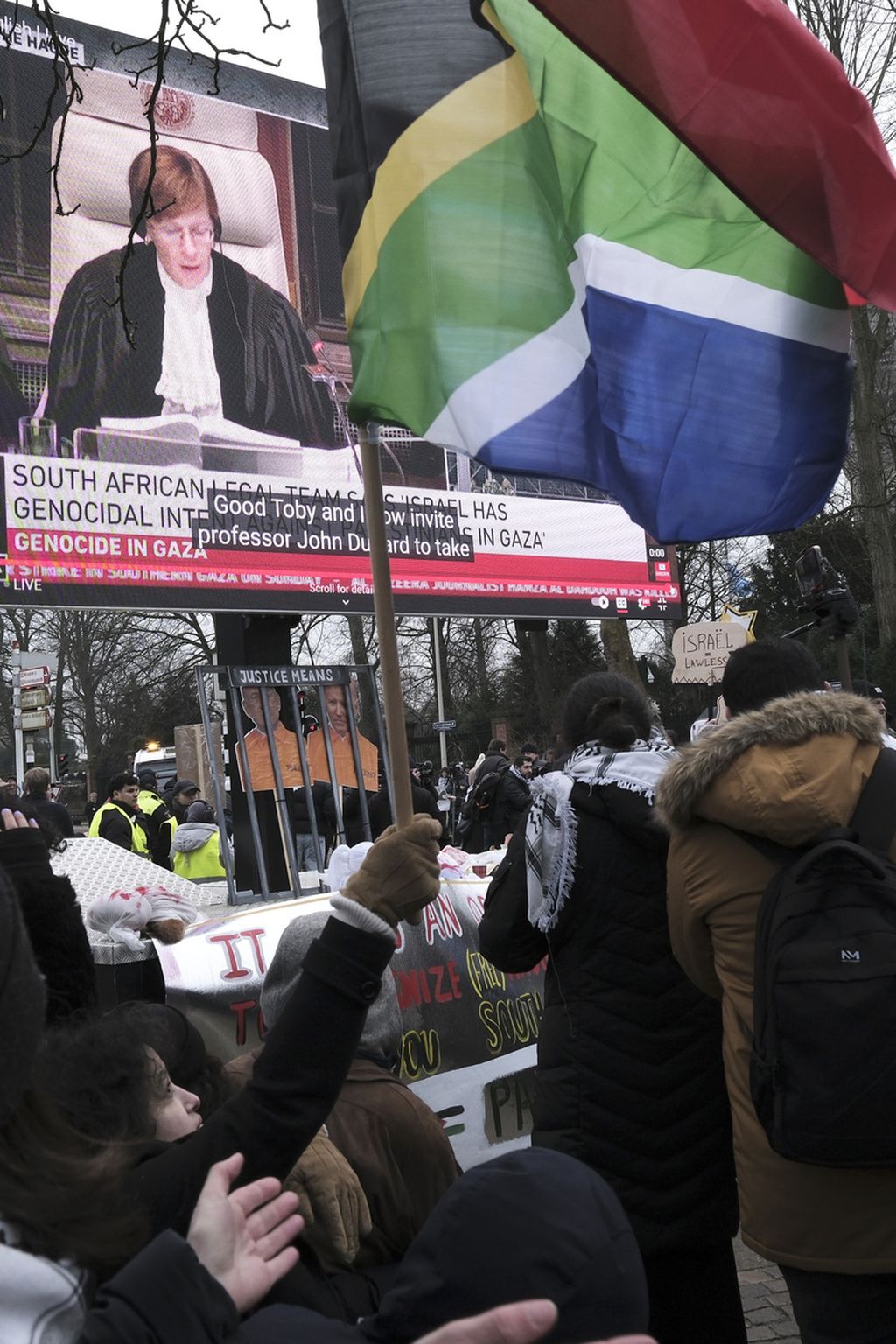Massa pengunjuk rasa melambaikan bendera Afrika Selatan di depan layar video raksasa di luar gedung Mahkamah Internasional (ICJ) di Den Haag, Belanda, sambil mendengarkan pernyataan pembukaan dalam gugatan Afrika Selatan yang mengajukan Israel melakukan genosida di Gaza, Kamis (11/1/2024). 