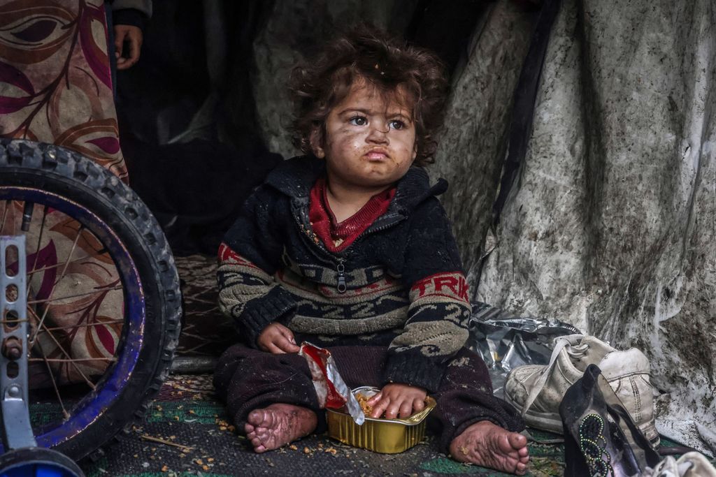 Seorang anak Palestina makan dari sebuah kotak sambil duduk di tenda pengungsi yang didirikan di tepi jalan di Rafah, Gaz, pada 14 Maret 2024. 