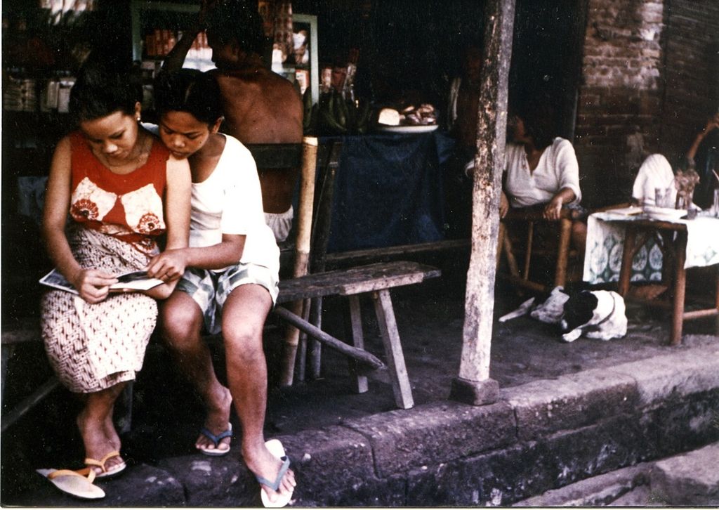 Ni Made Masih (berbaju merah, berkain) bersama Korni (berkaus putih) duduk di bangku panjang di bagian depan Warung Made, Kuta, Bali, tahun 1973. Keduanya sedang melihat foto-foto yang dihadiahkan seorang wisatawan. 