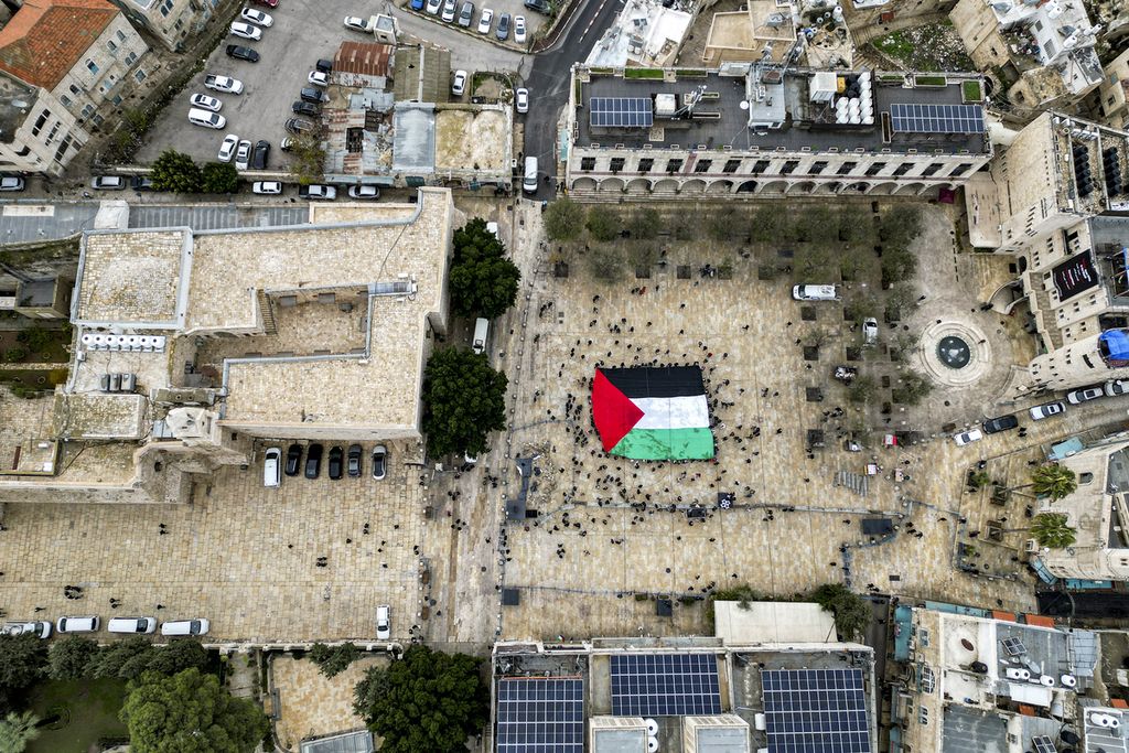 Pemandangan dari udara ini saat umat Krsitiani yang bersuka ria mengibarkan bendera raksasa Palestina di Manger Square di luar Gereja Kelahiran di kota Betlehem di Tepi Barat yang diduduki Israel, Minggu (24/12/2023). 