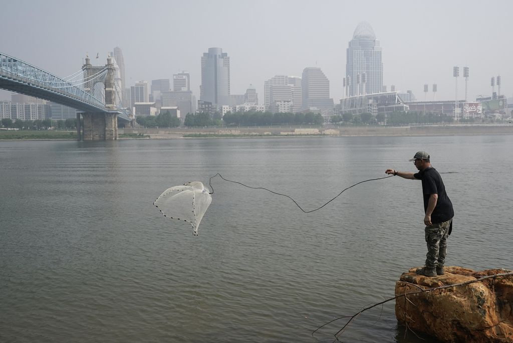 Seorang warga menangkap ikan menggunakan jaring di Sungai Ohio, Cincinnati, Rabu (28/6/2023), Kabut asap di latar belakang, yang menyelimuti kota Cincinnati terjadi karena "ekspor" asap dari Kanada. 