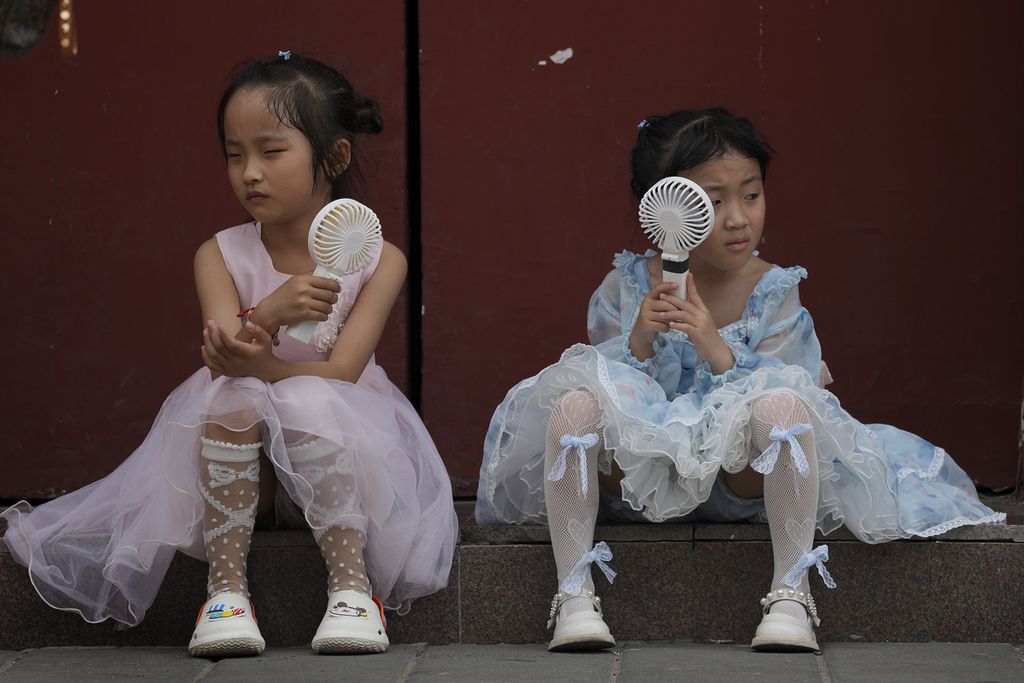Two children rest while holding an electric fan to ward off the heat during a break not far from the Forbidden City in Beijing, China, on Sunday (25/6/2023). In the past week, Beijing and several cities in northern China have been hit by scorching heat waves.