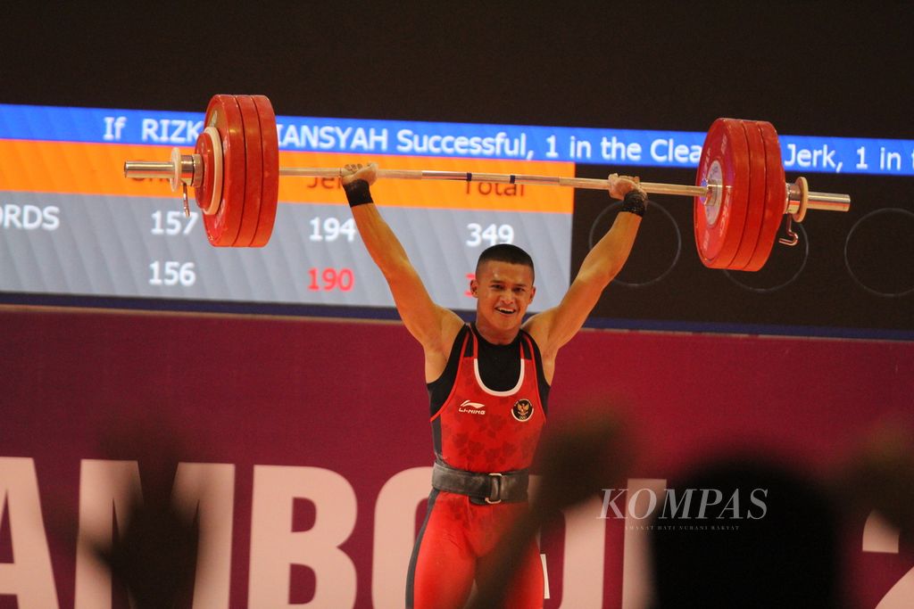 Dengan penuh percaya diri, Rizki Juniansyah, lifter muda Indonesia yang masih berusia 20 tahun, mengangkat pelat-pelat besi seusai memecahkan rekor dengan total angkatan 347 kilogram (<i>snatch </i>156 kg dan <i>clean and jerk</i> 347 kg) di cabang angkat besi kelas putra 73 kilogram di National Olympic Stadium, Kamboja, Minggu (14/5/2023).
