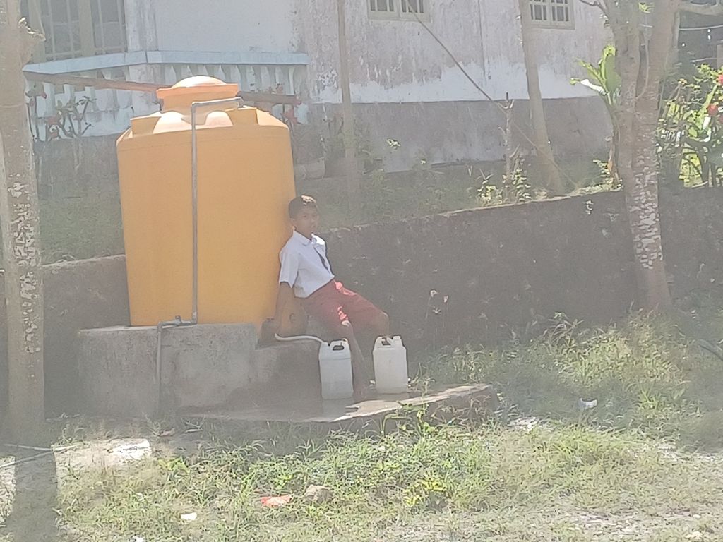 Hery Akoit (10), siswa SD Tunbaun, Kabupaten Kupang, 30 km arah utara Kota Kupang, Rabu (23/8/2023), duduk bersandar di tandon air ukuran 1.100 liter air, di samping gedung sekolahnya. Ia bersama sejumlah rekan mengambil air dari tandon itu untuk kebutuhan sekolah.