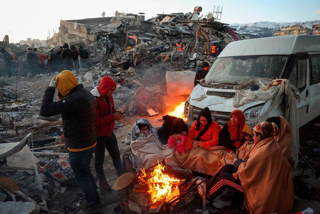 Para penyintas berkumpul di antara api unggun dengan latar belakang bangunan yang runtuh di Kahramanmaras, Turki, Rabu (8/2/2023). Gempa besar menewaskan lebih dari 17.000 orang di Turki dan diperkirakan jumlah korban akan terus meningkat. 
