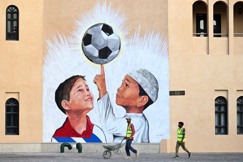 Seorang pekerja mendorong gerobak sorong sembari melewati sebuah mural di Doha, Selasa (8/11/2022), jelang turnamen sepak bola Piala Dunia FIFA 2022. 