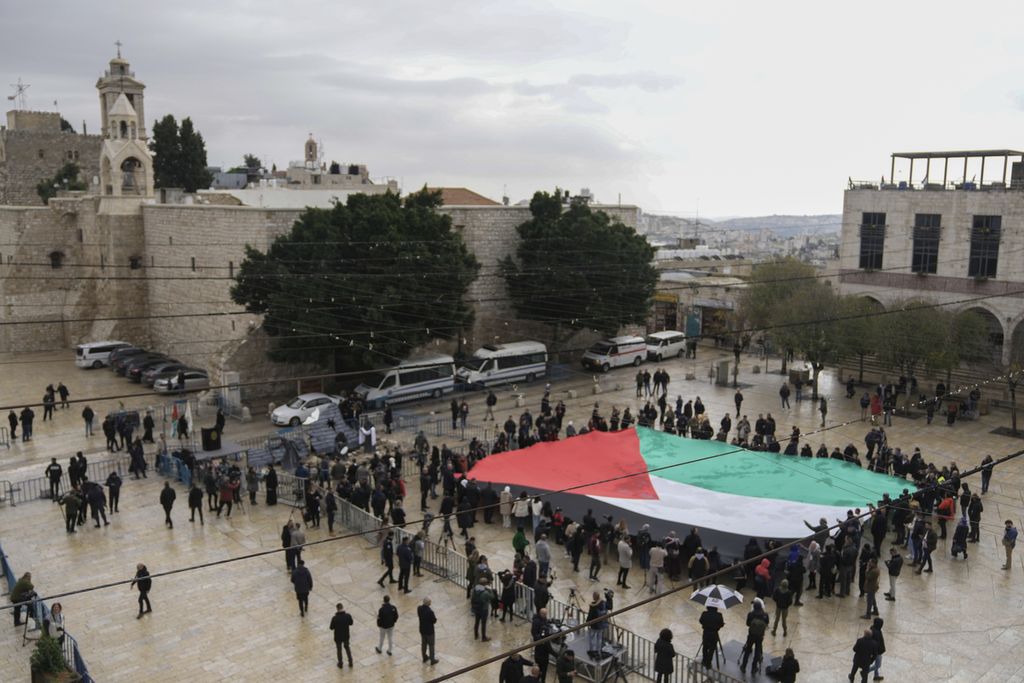 Orang-orang mengibarkan bendera raksasa Palestina di Manger Square, dekat Gereja Kelahiran, yang secara tradisional diyakini sebagai tempat kelahiran Yesus pada Malam Natal, di kota Bethlehem, Tepi Barat, Minggu (24/12/2023). 
