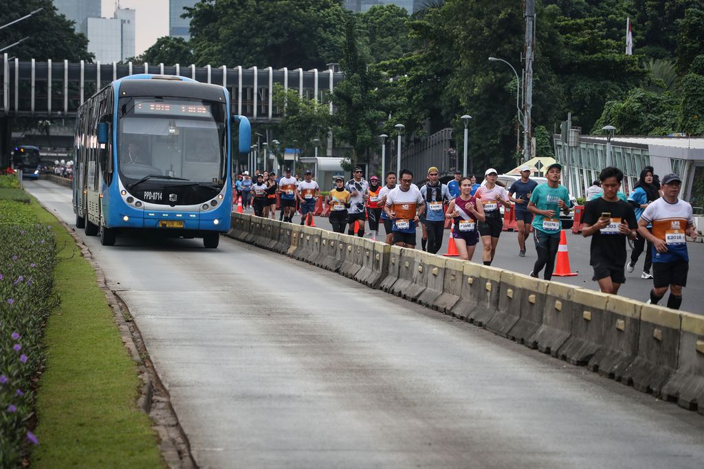 Peserta LPS Monas Half Marathon berlari di samping bus Transjakarta yang melintas di Jalan Jenderal Sudirman, Jakarta, Minggu (2/7/2023). Acara yang diikuti sekitar 5.000 pelari ini menjadi kesempatan bagi para pelari untuk menikmati Kota Jakarta sembari berolahraga. 