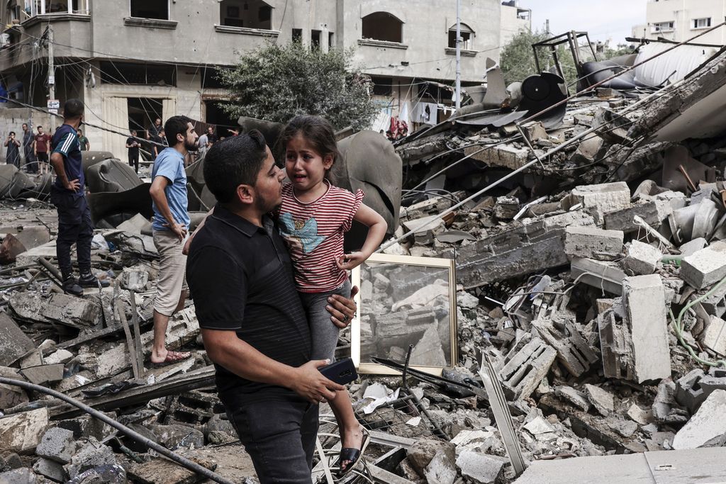 Seorang pria menggendong anak-anak yang sedang menangis saat berjalan di depan bangunan yang hancur akibat serangan udara Israel di Gaza, Sabtu (7/10/2023). 