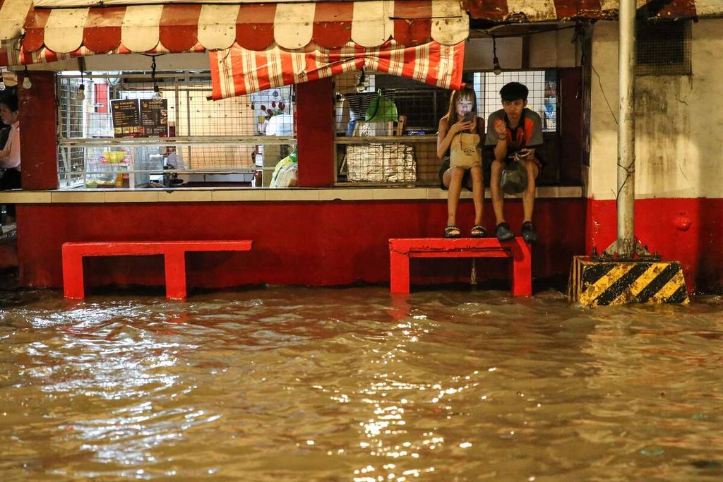 Dua orang warga Manila, Filipina tengah menunggu kendaraan umum di depan sebuah kedai sambil menghindari banjir yang menggenangi jalan raya. Foto diambil pada Sabtu 29/7/2023).