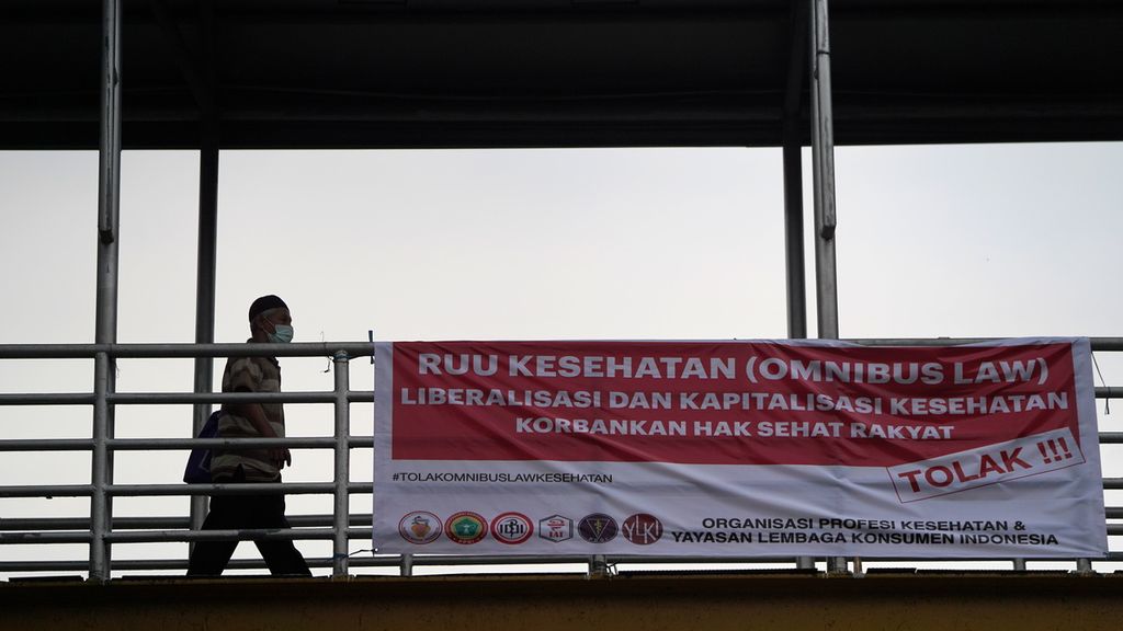 Warga melewati spanduk penolakan RUU Kesehatan di Tebet, Jakarta Selatan, Junat (25/11/2022). Rancangan Undang-Undang Kesehatan yang disusun dengan menghimpun sejumlah regulasi atau <i>omnibus law </i>masuk ke dalam Program Legislasi Nasional Prioritas tahun 2023. Namun, sampai saat ini penyusunan RUU ini tidak pernah melibatkan organisasi profesi kedokteran. 