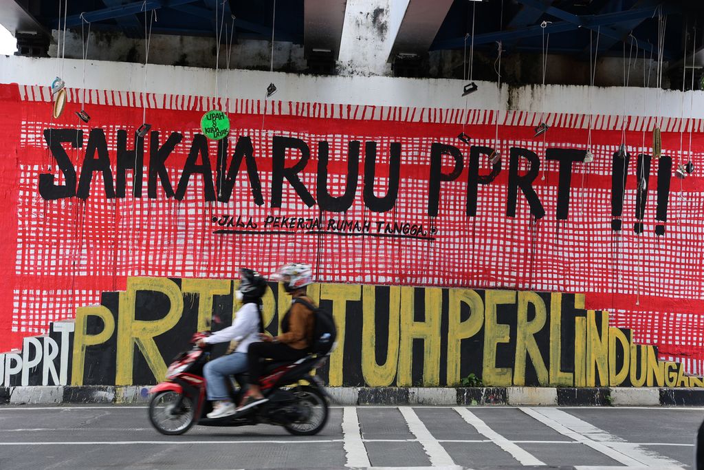 Pengguna sepeda motor melintas di depan mural berisi kritik tentang pembahasan RUU Perlindungan Pekerja Rumah Tangga yang tidak kunjung selesai di Jembatan Kewek, Yogyakarta, akhir Desember 2021. 
