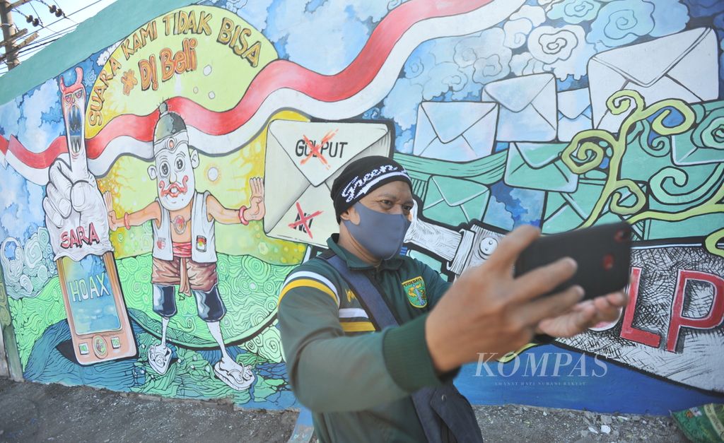 Warga berfoto di depan mural bertema Pilkada Serentak di Jalan Raya Ahmad Yani, Kecamatan Waru, Sidoarjo, Jawa Timur, Senin (24/8/2020). Mural mengajak masyarakat untuk aktif berpartisipasi dan menolak segala bentuk politik uang dan hoaks pada pilkada.