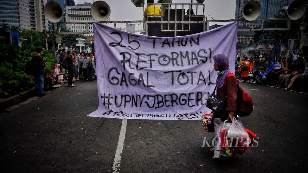 Pedagang asongan melintasi spanduk aksi mahasiswa dari sejumlah perguruan tinggi di Jabodetabek ketika menggelar Aksi Peringatan 25 Tahun Reformasi di kawasan Patung Kuda, Jalan Merdeka Barat, Jakarta, Kamis (25/5/2023). 
