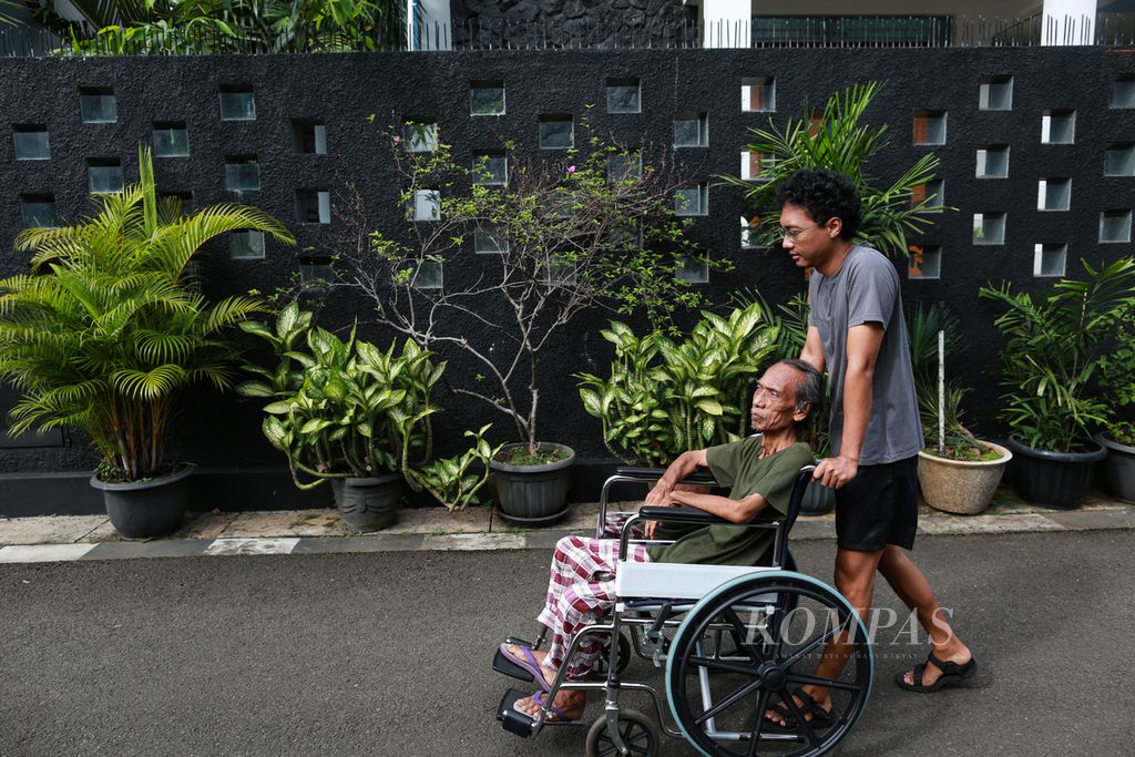 Sri Warso Wahono (74) duduk di kursi roda ditemani putranya, Aria, mengelilingi kompleks untuk menikmati sinar matahari pagi di kawasan Palmerah, Jakarta Barat, Rabu (25/5/2022). Keliling komplek setiap pagi menjadi rutinitas yang mereka lakukan bersama.