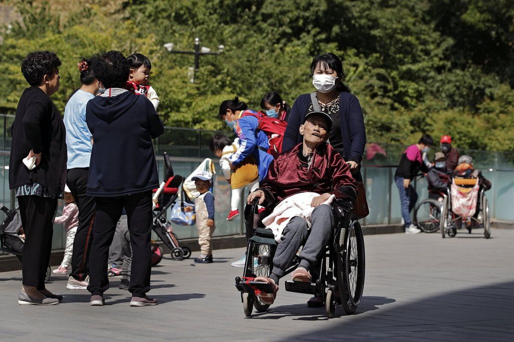 Seorang wanita yang memakai masker wajah mendorong seorang pria lanjut usia di kursi roda berjalan-jalan di taman di dekat gedung perkantoran komersial di Beijing, China, Senin (10/5/2021). AP/ANDY WONG