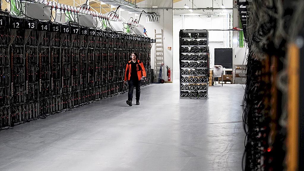 Dalam  foto bertanggal 17 Januari 2018 ini, seorang pekerja berjalan di antara deretan komputer yang melakukan penambangan bitcoin, di  pusat penambangan mata uang virtual  Genesis Mining, di Keflavik, Eslandia. Dibutuhkan banyak energi listrik agar komputer-komputer dapat beroperasi untuk menambang bitcoin.
