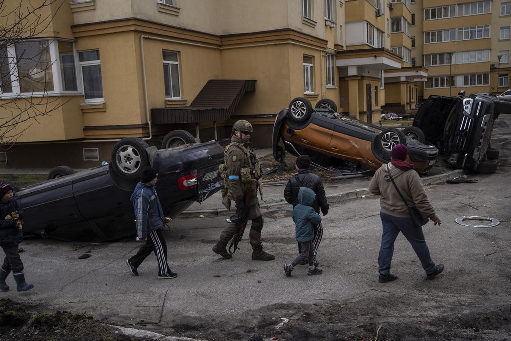 Seorang tentara Ukraina berjalan bersama bocah-bocah melewati mobil-mobil yang terbalik dan hancur dalam perang di Bucha, Ukraina, Senin (4/4/2022). Otoritas setempat mengatakan kepada The Associated Press bahwa setidaknya 16 anak-anak termasuk dalam ratusan korban tewas di Bucha. 