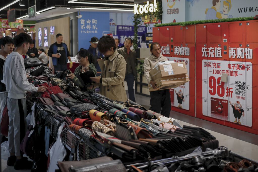 A worker carrying boxes of goods walks by women selecting handbags at a commercial building in Beijing, Friday, Oct. 18, 2019. China's economic growth sank to a 26-year low in the latest quarter amid pressure from a trade war with Washington, adding to a deepening slump that is weighing on global growth. 