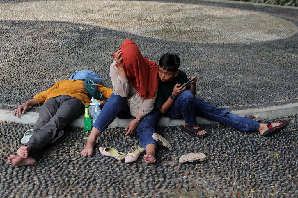 Anak bermain gawai di samping orangtuanya yang tertidur di Taman Lapangan Banteng, Jakarta Pusat, Minggu (2/7/2023). 